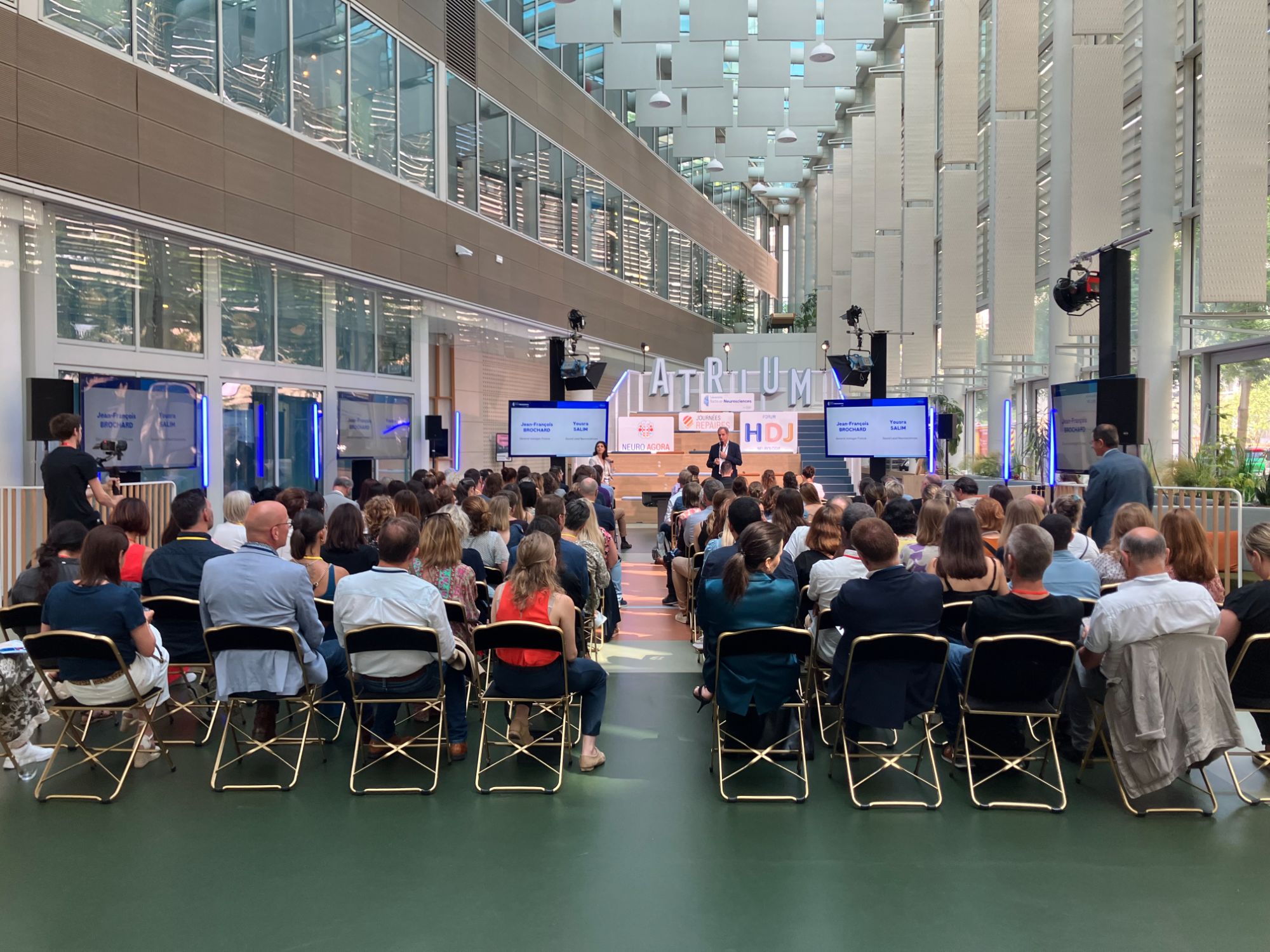 Conférence en atrium : salle assise, scène éclairée, écrans de retransmission de part et d’autre et enceintes sur pied